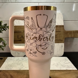 May include: A pink stainless steel travel mug with a gold lid and handle. The mug features a design of a stethoscope, flowers, and medical tools. The name "Kimberly" is written in gold lettering on the mug.