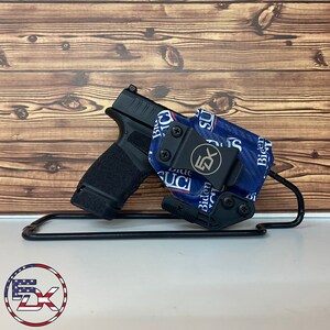 May include: A black handgun with a textured grip is displayed with a blue and white patterned holster. The holster features the text "Biden Sucks" and a logo. The setup is presented on a black metal stand against a wooden background.