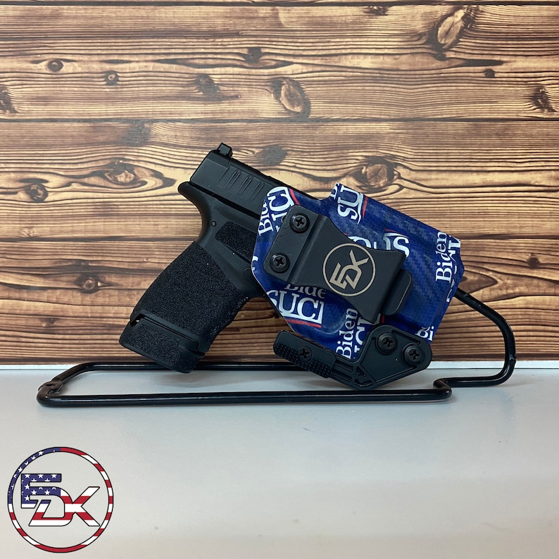 May include: A black handgun with a textured grip is displayed with a blue and white patterned holster. The holster features the text "Biden Sucks" and a logo. The setup is presented on a black metal stand against a wooden background.