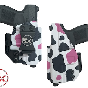 May include: A black and pink cow print gun holster with a black clip. The holster is designed to fit a handgun with a black polymer frame.