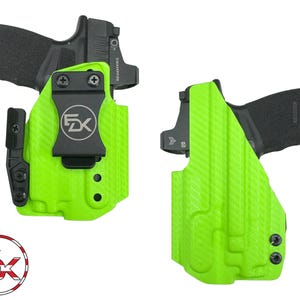May include: A bright neon green molded holster containing a black semi-automatic pistol with an optic sight. The holster features a black belt clip with a circular logo reading FDK. A matching circular logo with a flag pattern is also visible.