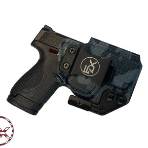May include: Black handgun with a black and blue camouflage holster. The holster has a black clip with the letters "5X" on it. The image is on a white background.