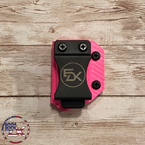 May include: A pink and black gun holster with a carbon fiber pattern and the text "FDK" in gold. The holster has a clip on the back and is designed to be worn on a belt.