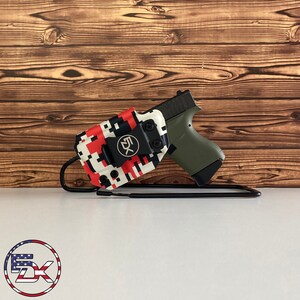 May include: A black handgun in an olive green grip is displayed in a red, white, and black pixelated holster. The holster has a black clip with the letters "5X" on it. The setup is on a white surface with a wooden background.