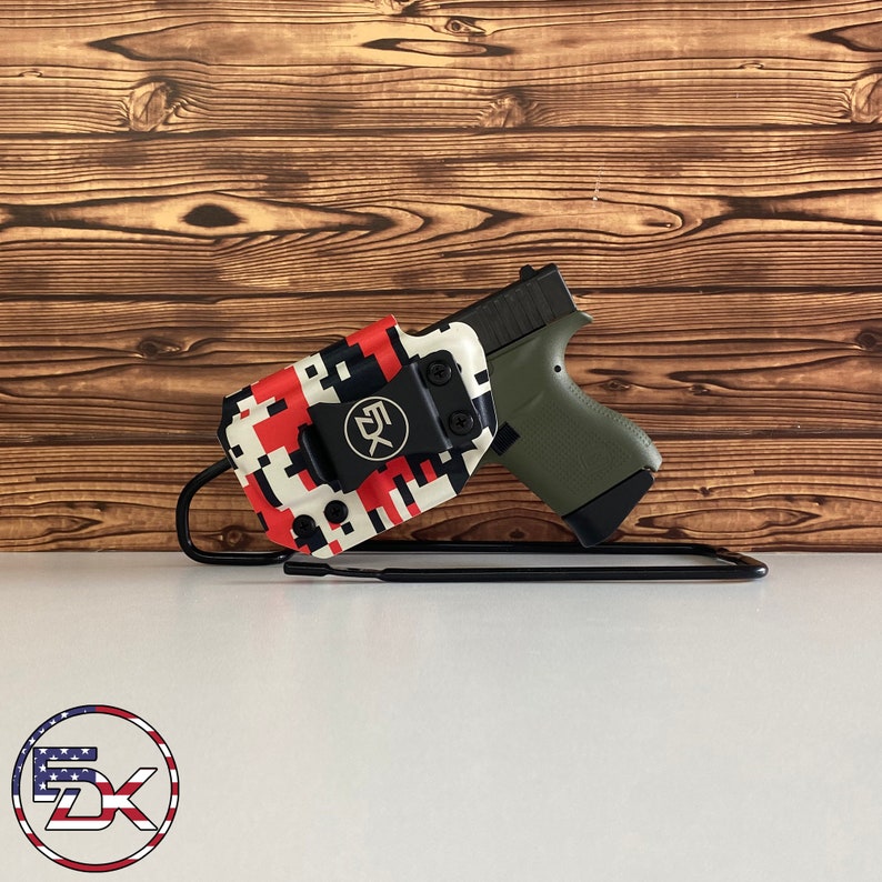 May include: A black handgun in an olive green grip is displayed in a red, white, and black pixelated holster. The holster has a black clip with the letters "5X" on it. The setup is on a white surface with a wooden background.