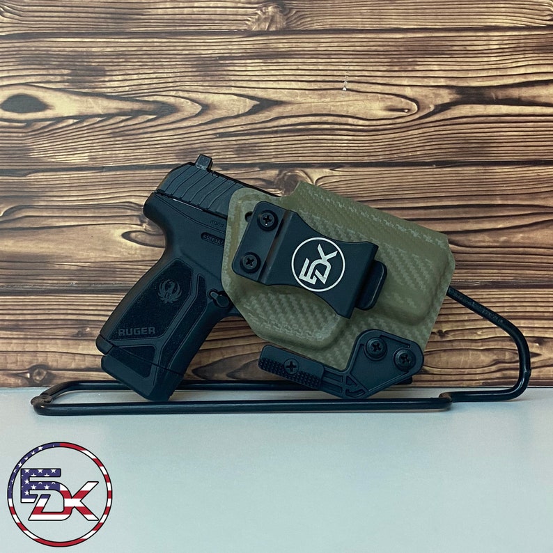 May include: A black Ruger pistol with a green carbon fiber holster. The holster has a black clip and a black logo that says "5DX". The pistol and holster are sitting on a black stand.