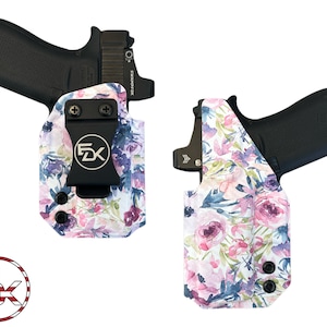 May include: A white holster with a floral pattern in shades of pink, purple, and blue. The holster has a black clip and is designed to hold a handgun. The holster is made by the company "FDX".