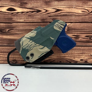 May include: A camouflage-patterned holster with a blue handgun inside. The holster is attached to a black metal hook.