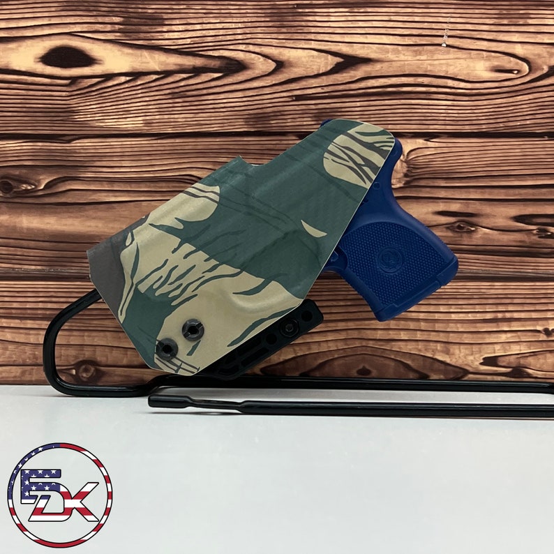 May include: A camouflage-patterned holster with a blue handgun inside. The holster is attached to a black metal hook.