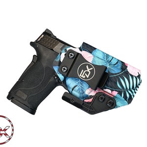 Puede incluir: Una pistola negra con una funda negra con estampado floral tropical con flores y hojas azules y rosas. La funda tiene un clip negro y un logotipo plateado que dice "EDX".