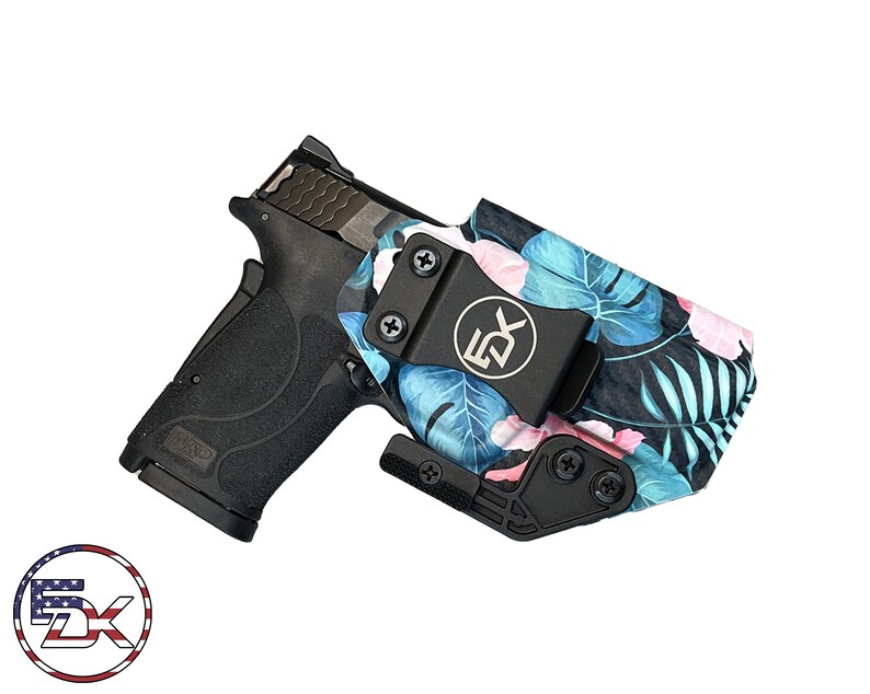 Puede incluir: Una pistola negra con una funda negra con estampado floral tropical con flores y hojas azules y rosas. La funda tiene un clip negro y un logotipo plateado que dice "EDX".