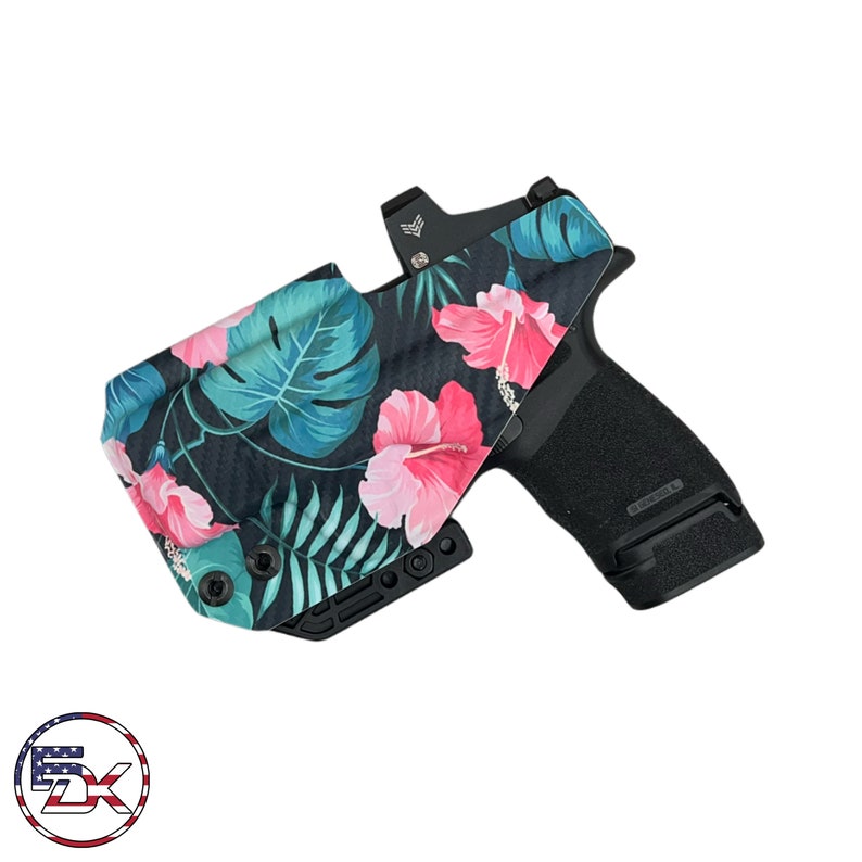 Puede incluir: Una funda para pistola de color negro y verde con estampado floral, con una pistola negra en su interior. La funda tiene un patr&oacute;n de fibra de carbono y presenta flores de hibisco rosas y hojas tropicales verdes.