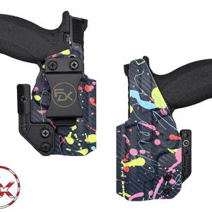 May include: Two black handguns in holsters. The holsters have a black carbon fiber pattern with colorful paint splatters in pink, yellow, and blue. The holsters have black clips and hardware. The logo "FX" is visible on the holster.