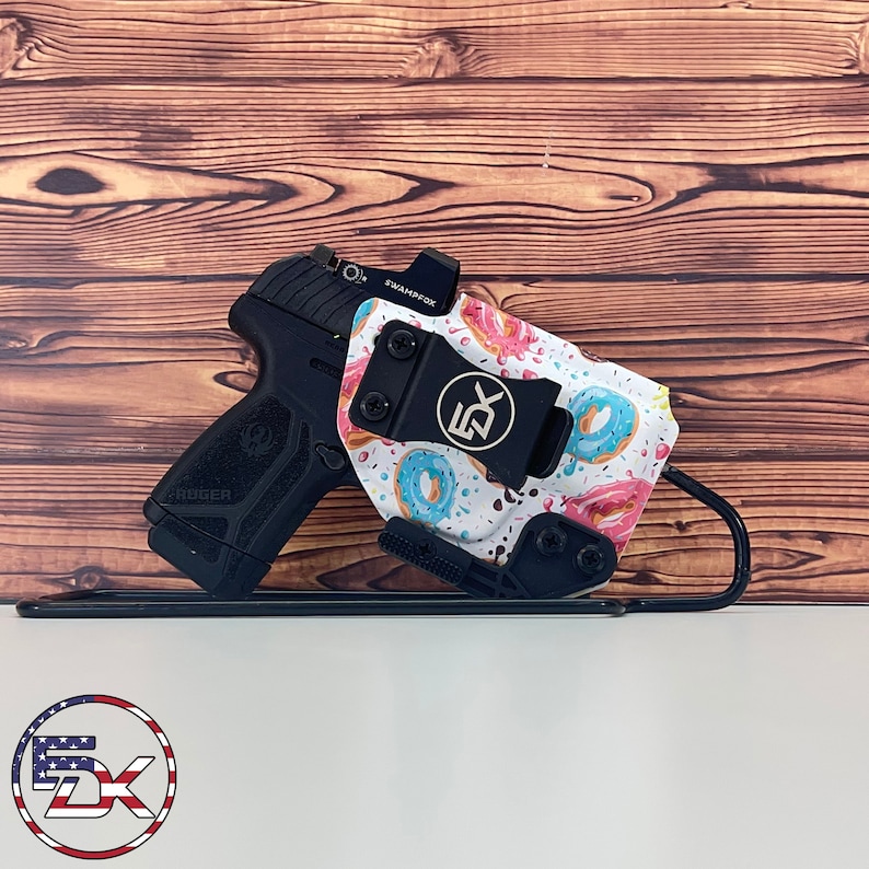 May include: Black handgun with a red dot sight and a holster featuring a white background with a pattern of donuts in pink, blue, and brown. The holster has a black clip and the logo "DX".