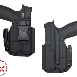 May include: Two black gun holsters are displayed against a white background. One holster has a gun inside, and the other is empty. The holsters have a clip with the letters "EK" inside a circle. The logo is red, white, and blue.