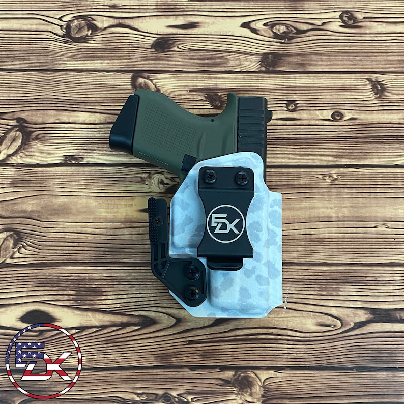 May include: An olive green handgun in a white and gray camouflage pattern holster with a black clip. The holster has a black logo with the letters "EK" inside a circle. The holster is on a wooden surface.