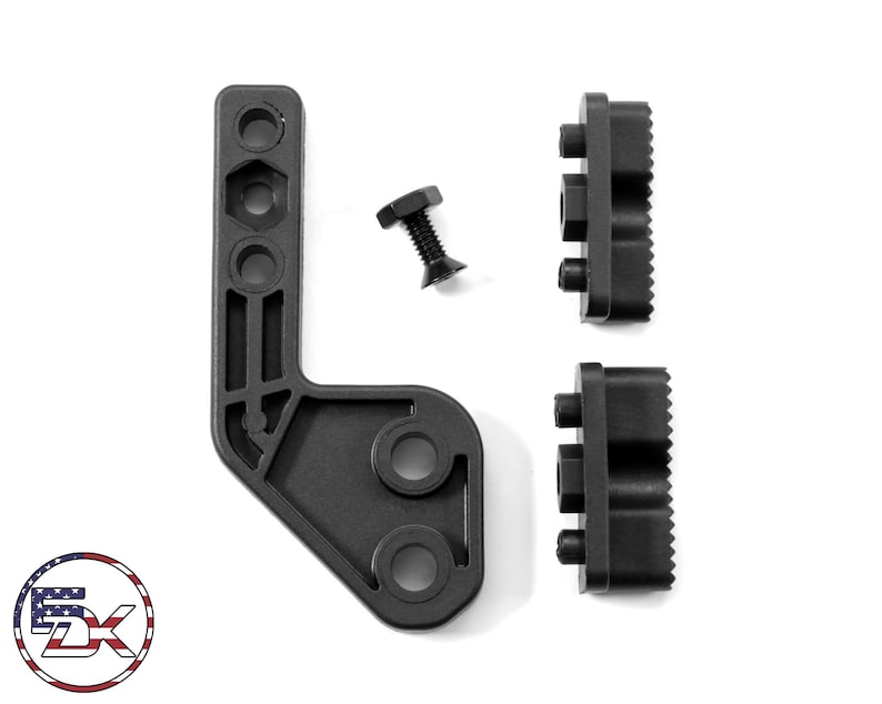 Op de afbeelding: Zwarte montagehardware set, inclusief een L-vormige beugel met meerdere gaten, twee rechthoekige stukken met ribbels en een schroef. De componenten zijn op een witte achtergrond geplaatst.