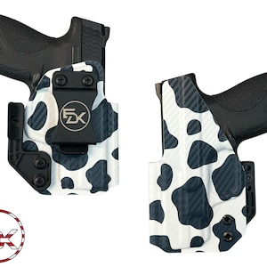 May include: Two black handguns are displayed in custom holsters. The holsters feature a white and black cow print design. The holsters have a black clip and hardware. The logo "5X" is visible in a circular design.