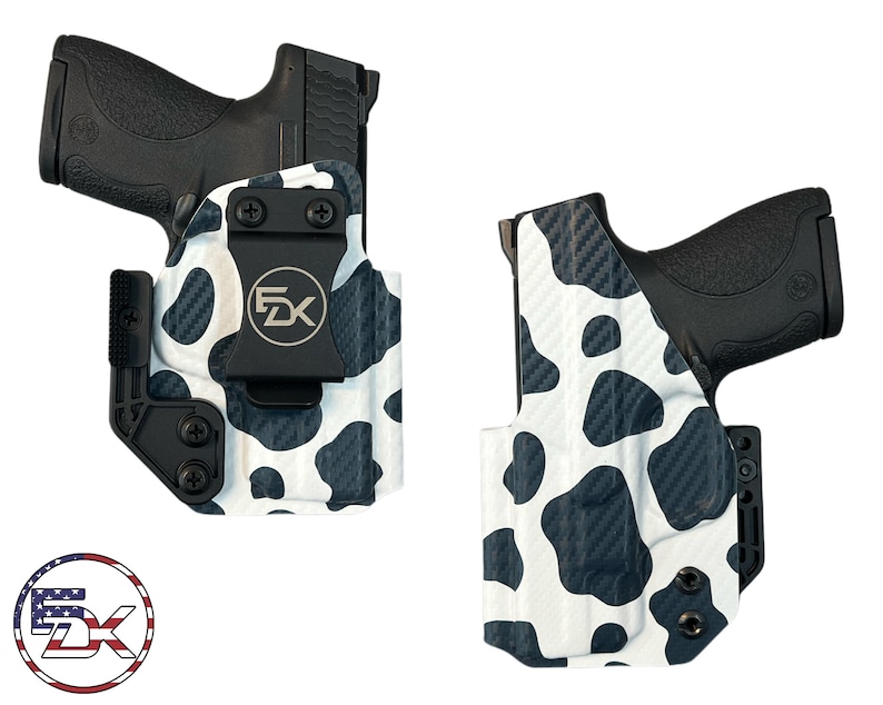 May include: Two black handguns are displayed in custom holsters. The holsters feature a white base with a black cow print pattern. The holsters have a black clip and hardware. The logo "5X" is visible in a circular design.