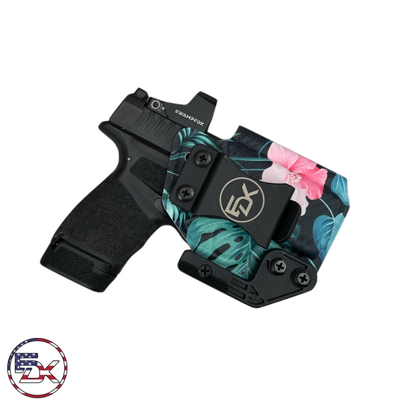 Puede incluir: Una pistola negra con una mira de punto rojo est&aacute; enfundada en una funda negra y verde con estampado floral tropical. La funda tiene un clip negro y un logotipo plateado que dice "EDX".