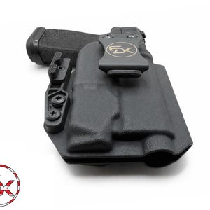 May include: Black polymer holster with a firearm inside. The holster has a belt clip with the letters "FX" and a logo. The firearm's grip is textured. The holster is designed for concealed carry.