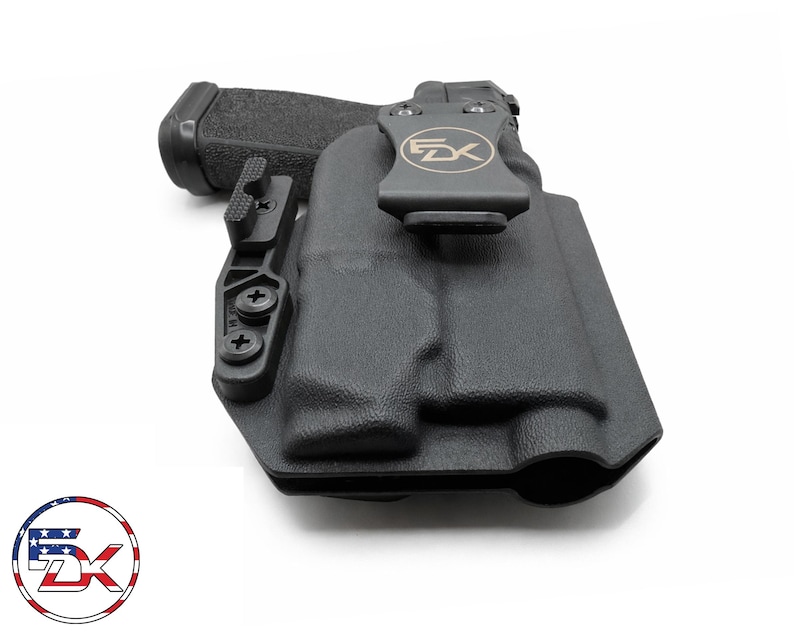 May include: Black polymer holster with a firearm inside. The holster has a belt clip with the letters "FX" and a logo. The firearm's grip is textured. The holster is designed for concealed carry.