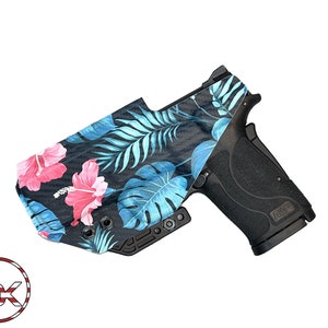 Puede incluir: Una pistola negra con una funda de estampado floral. La funda presenta un estampado tropical con flores de hibisco rosas y hojas azules.