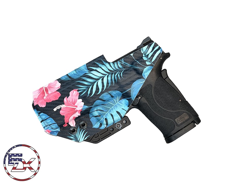 Puede incluir: Una pistola negra con una funda de estampado floral. La funda presenta un estampado tropical con flores de hibisco rosas y hojas azules.