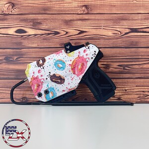 May include: A black handgun with a white holster decorated with a colourful donut pattern. The donuts are pink, brown, and blue with sprinkles. The gun rests on a black stand against a wooden backdrop.