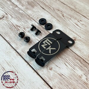 May include: A black metal bracket with the letters "EK" inside a circle, along with screws, washers, and other hardware. The items are arranged on a light wood grain surface. The logo of "5X" is in the bottom left corner.