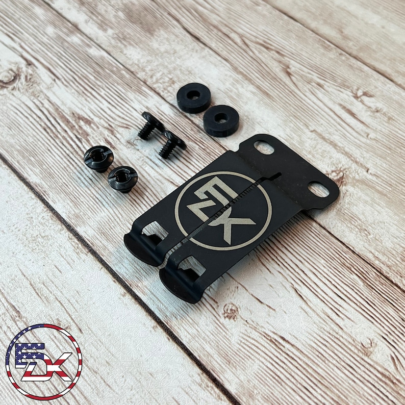 May include: A black metal bracket with the letters "EK" inside a circle, along with screws, washers, and other hardware. The items are arranged on a light wood grain surface. The logo of "5X" is in the bottom left corner.