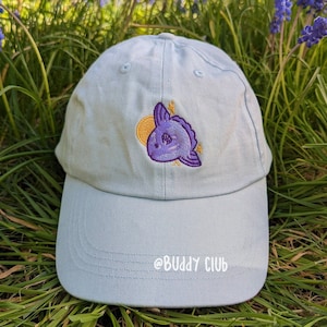 May include: A light blue baseball cap with an embroidered purple and yellow ocean sunfish design. The text "@BUDDY CLUB" is embroidered on the brim of the cap.