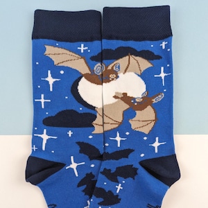 May include: Blue socks with a bat and moon design. The socks feature a bat illustration with a moon and stars, and the text "Buddy Club" at the cuff. The socks have a dark blue cuff and toe.