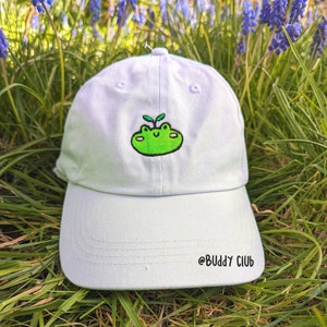 May include: A light blue baseball cap with an embroidered green frog with a sprout on its head. The cap has the text "@BUDDY CLUB" embroidered on the brim.