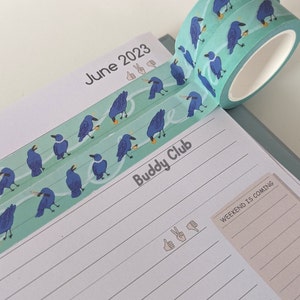 Puede incluir: Un rollo de cinta washi turquesa con un patrón repetitivo de pájaros azules que sostienen un pequeño objeto amarillo. La cinta está parcialmente desenrollada y colocada en una página de un planificador con el texto "June 2023" y "Buddy Club" escrito en ella.