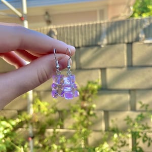K&ouml;nnte beinhalten: Ein Paar silberne Ohrringe mit einem durchsichtigen lila Gummib&auml;rchen-Anh&auml;nger.