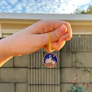 May include: A gold chain necklace with a round pendant featuring a blue and white enamel depiction of Hogwarts castle with stars and a crescent moon. The pendant reads "HOGWARTS" in gold lettering.
