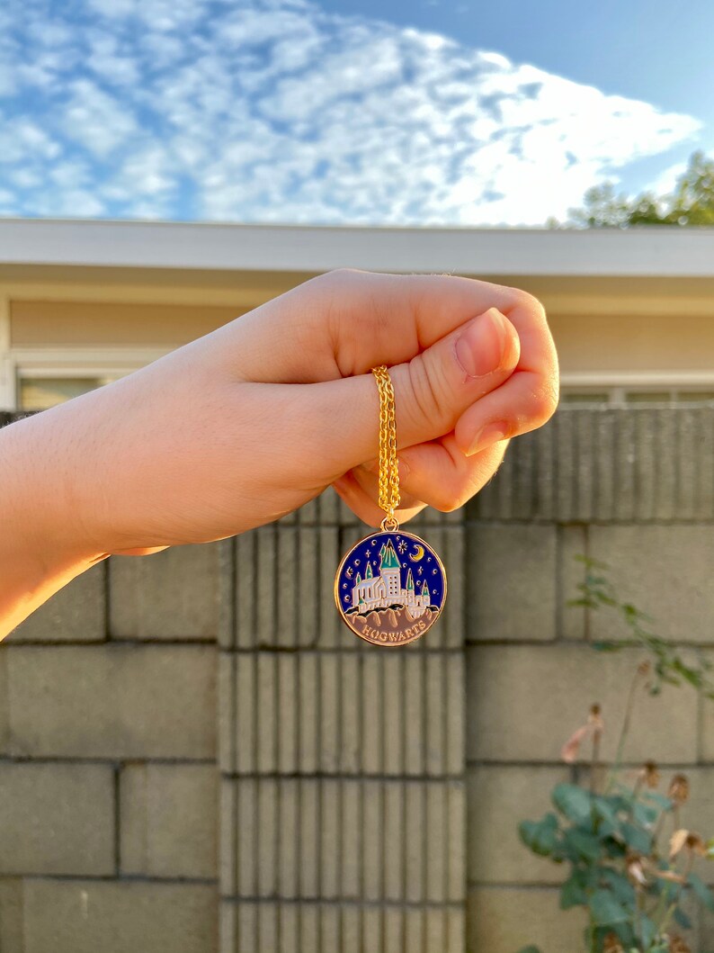 May include: A gold chain necklace with a round pendant featuring a blue and white enamel depiction of Hogwarts castle with stars and a crescent moon. The pendant reads "HOGWARTS" in gold lettering.