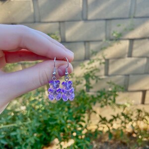 K&ouml;nnte beinhalten: Ein Paar silberne Ohrringe mit violetten Gummib&auml;rchen-Anh&auml;ngern.