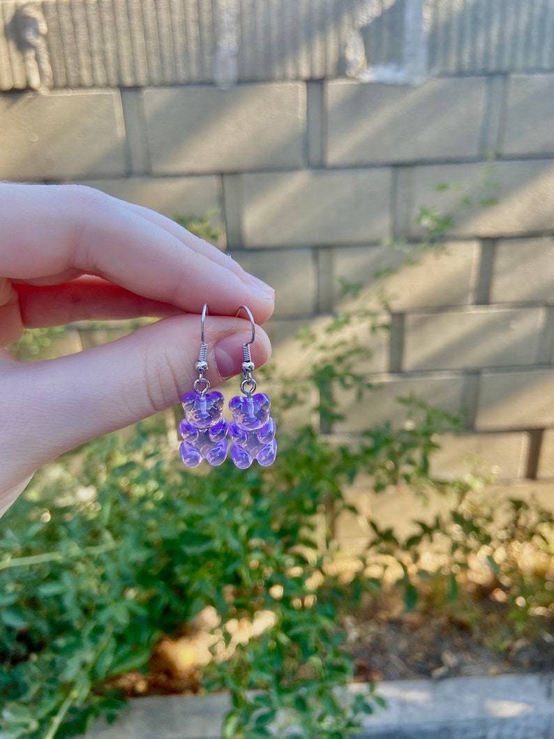 K&ouml;nnte beinhalten: Ein Paar silberne Ohrringe mit violetten Gummib&auml;rchen-Anh&auml;ngern.