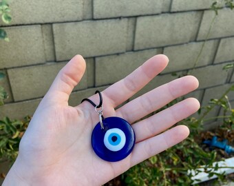 May include: A blue glass evil eye pendant necklace with a black cord. The pendant is round and has a white center with a blue eye.