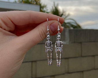 Puede incluir: Un par de pendientes de esqueleto de plata. Los pendientes están hechos de metal y presentan un diseño de esqueleto detallado.