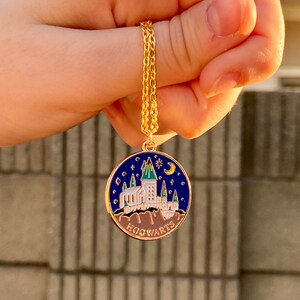 May include: A gold chain necklace with a round pendant featuring a blue enamel Hogwarts castle design. The pendant has a white castle with green accents and a crescent moon and star on top. The text "HOGWARTS" is written in gold at the bottom of the pendant.