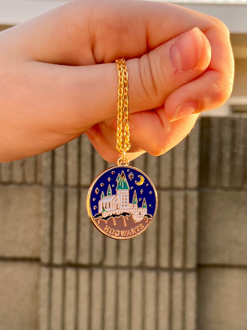 May include: A gold chain necklace with a round pendant featuring a blue enamel Hogwarts castle design. The pendant has a white castle with green accents and a crescent moon and star on top. The text "HOGWARTS" is written in gold at the bottom of the pendant.