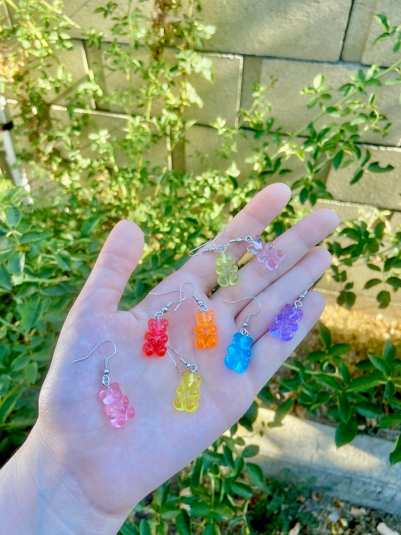 K&ouml;nnte beinhalten: Regenbogen-Gummib&auml;rchen-Ohrringe zum H&auml;ngen. Die Ohrringe sind aus klarem Kunststoff mit einem Gummib&auml;rchen-Anh&auml;nger in jeder Farbe des Regenbogens: Rot, Orange, Gelb, Gr&uuml;n, Blau und Lila.