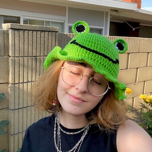 Puede incluir: Un sombrero de cubo de rana verde tejido a crochet con ojos negros y una boca sonriente. El sombrero lo lleva una persona con cabello castaño y gafas.