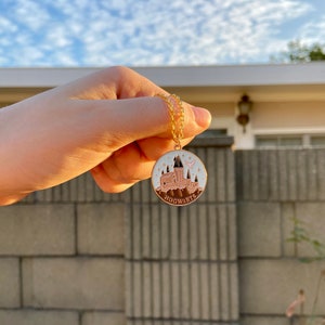 May include: A gold chain necklace with a round pendant featuring a pink and white illustration of Hogwarts castle with the word "HOGWARTS" printed below.