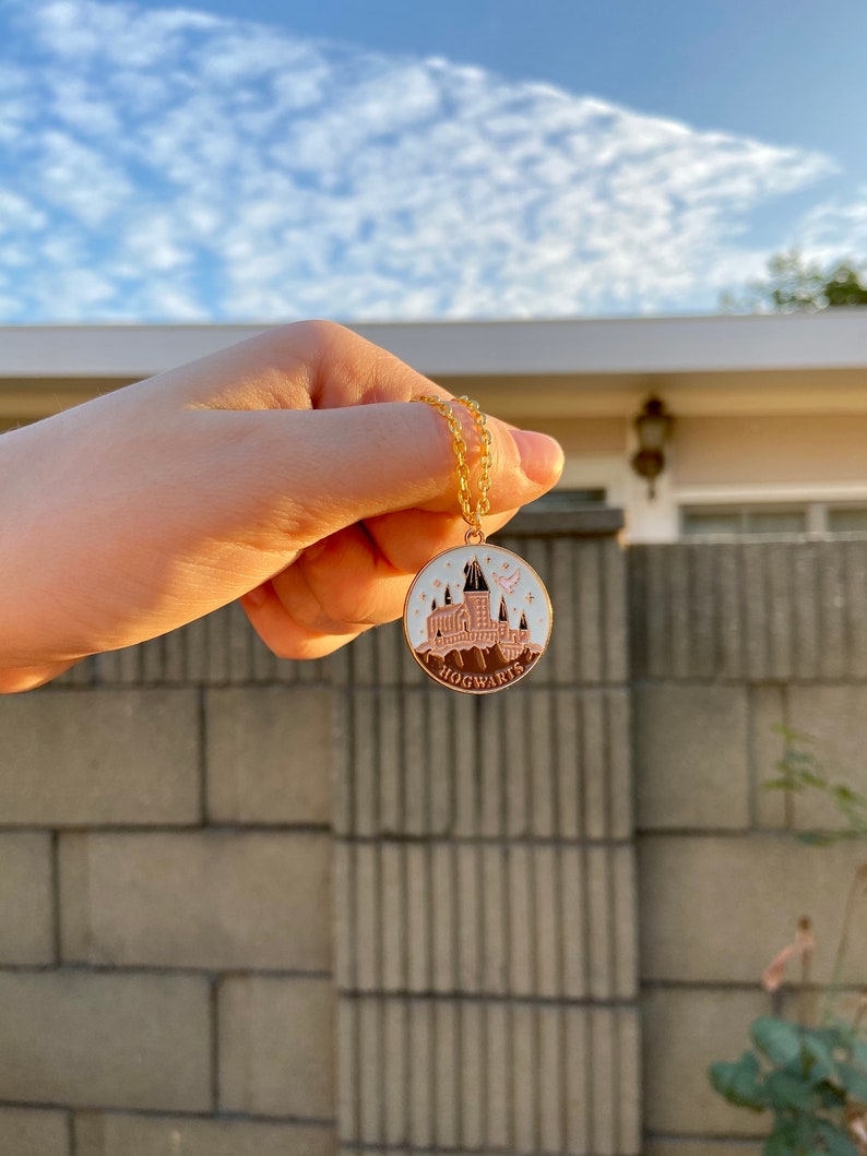 May include: A gold chain necklace with a round pendant featuring a pink and white illustration of Hogwarts castle with the word "HOGWARTS" printed below.