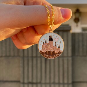 May include: A gold chain necklace with a round pendant featuring a white enamel depiction of Hogwarts castle with a pink bird flying above. The pendant has a brown enamel border and the word "HOGWARTS" written in brown enamel.