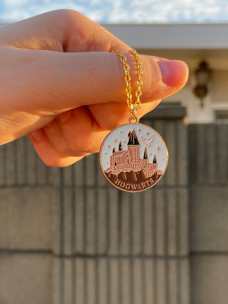 May include: A gold chain necklace with a round pendant featuring a white enamel depiction of Hogwarts castle with a pink bird flying above. The pendant has a brown enamel border and the word "HOGWARTS" written in brown enamel.